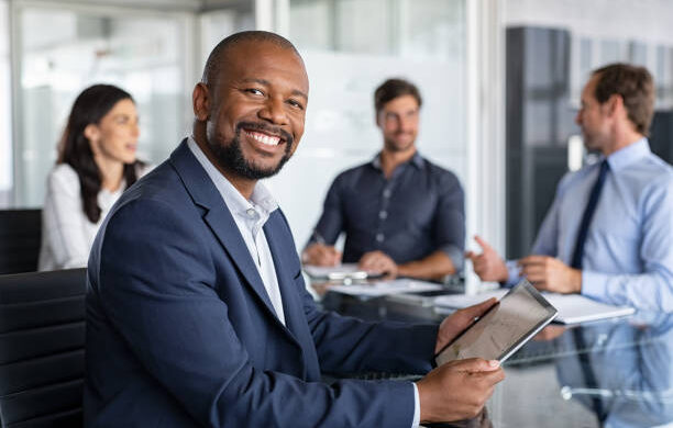 Mature black businessman with colleagues sitting in a modern board room. Proud smiling business man sitting during a meeting and looking at camera. Portrait of happy successful executive with team working in background.