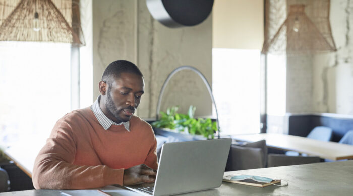 Portrait of adult African-American man using laptop while working in modern cafe interior decorated with fresh green plants, copy space