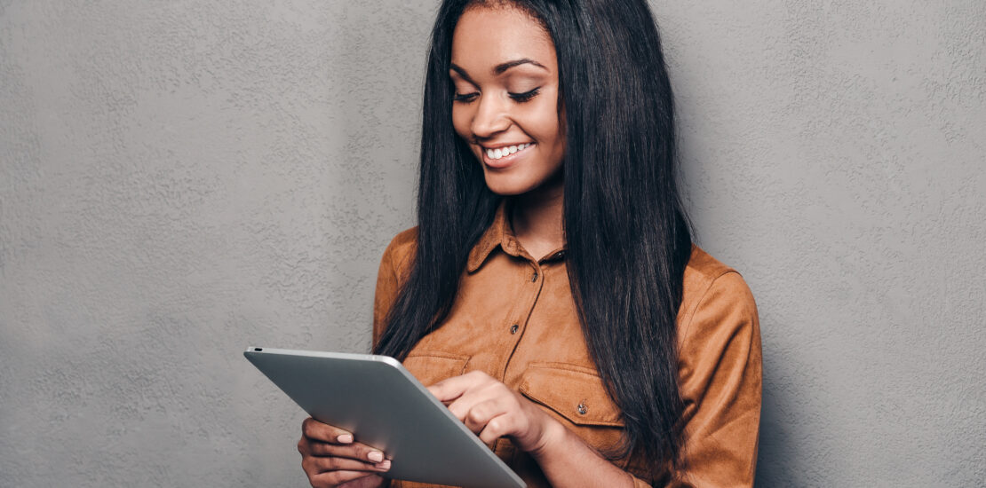 Beauty With Digital Tablet. Beautiful Young African Woman Holding Touchpad And Looking At It With Smile While Standing Against Grey Background