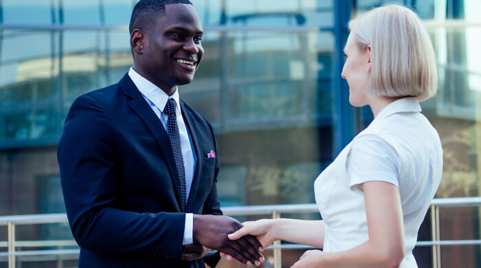 handsome African American man in a black business suit, shaking hand with a businesswoman partner cityscape glass offices background. teamwork and successful deal idea