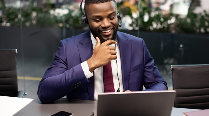 Handsome young black manager in expensive suit having video interview with employee, looking at laptop screen and smiling, using headset, modern office interior, copy space