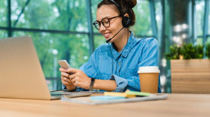 Smiling confident businesswoman checking her phone with headset and laptop at office desk