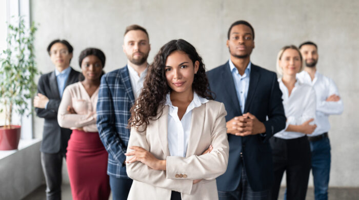 Successful Businesswoman Standing In Front Of Business Team In Office Successful Latin Businesswoman Standing In Front Of Business Team Posing In Modern Office, Smiling To Camera. Female Entrepreneurship And Leadership, Career Growth And Success. Selective Focus