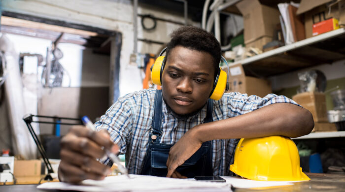 Serious thoughtful handsome young African-American construction engineer in soundproof headphone sitting at table and making notes in diary while planning work in dark room