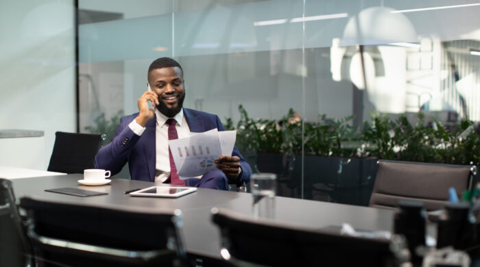Wealthy black guy in suit entrepreneur having phone conversation with his assistant or business partner, sitting at table with digital tablet on, looking at documents, panorama with copy space