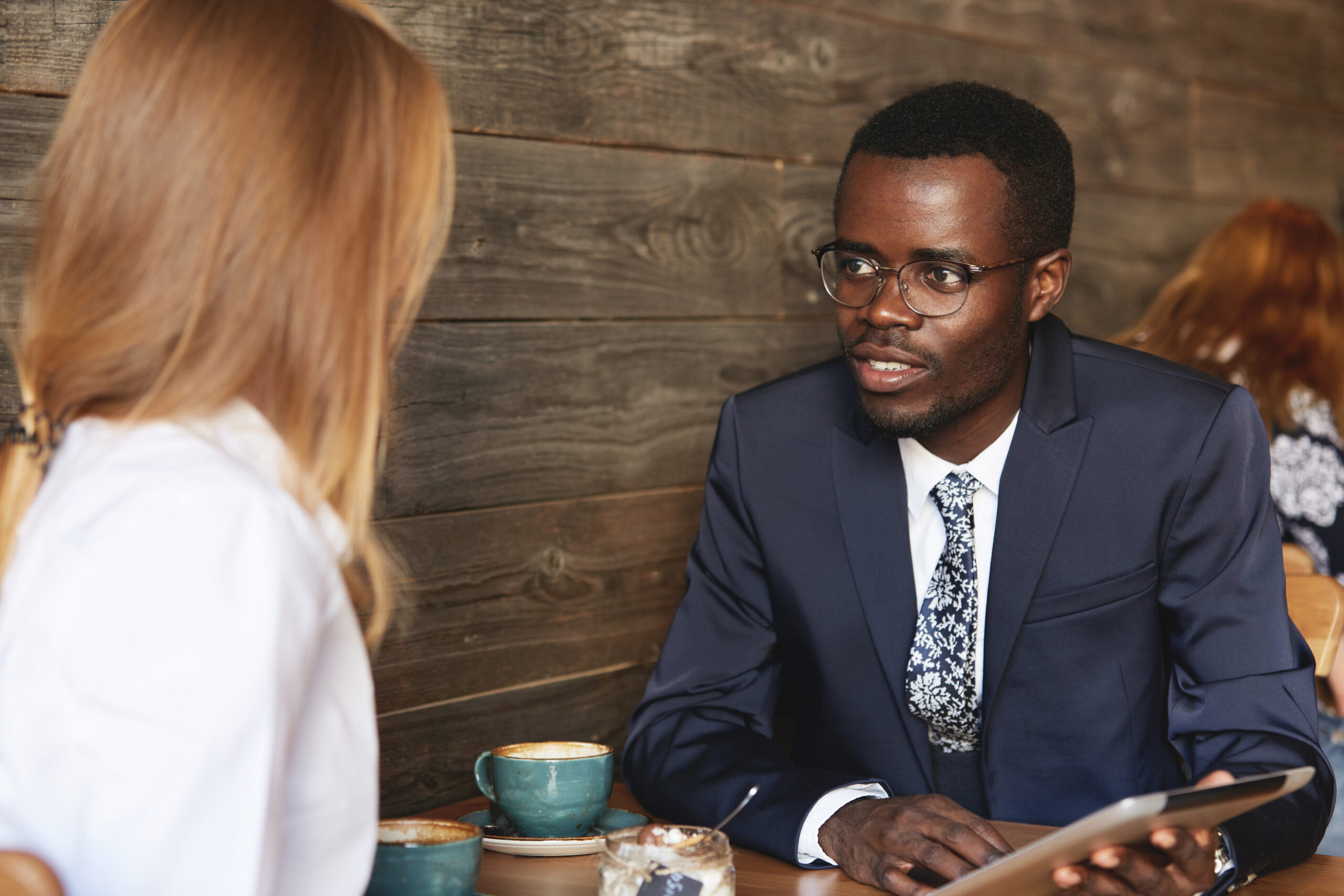 Young African Businessman Holding Tablet And Sharing Data With Female Caucasian Colleague During Coffee Break In Cosy Caf�smart Coworker Makes An Eye Contact With Job Partner And Listens To Her.