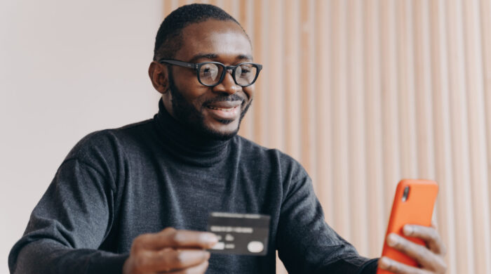 Young happy african american male entrepreneur with credit card and mobile phone using e-banking app, smiling afro man in glasses buying good in internet store, making financial transactions online