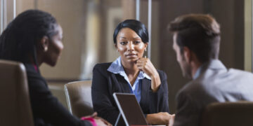 A group of three multi-ethnic businesspeople in a meeting in an office boardroom.  They are sitting, and there is a digital tablet on the table.  The focus is on the young African American woman with her hand on her chin, wearing a black suit.