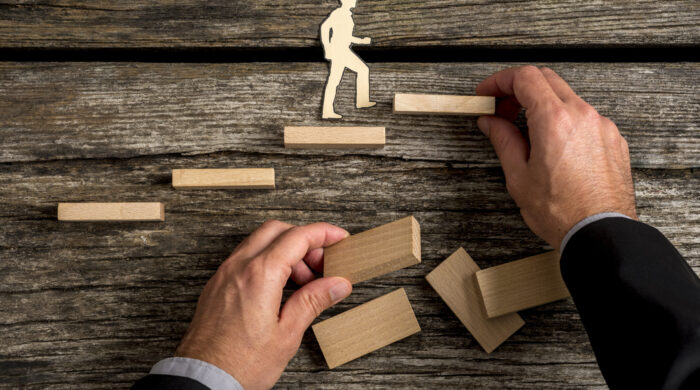 Silhouette cutouts of a man walking up steps, while the hand of other businessman building stairs for him in a conceptual image of personal and career promotion.
