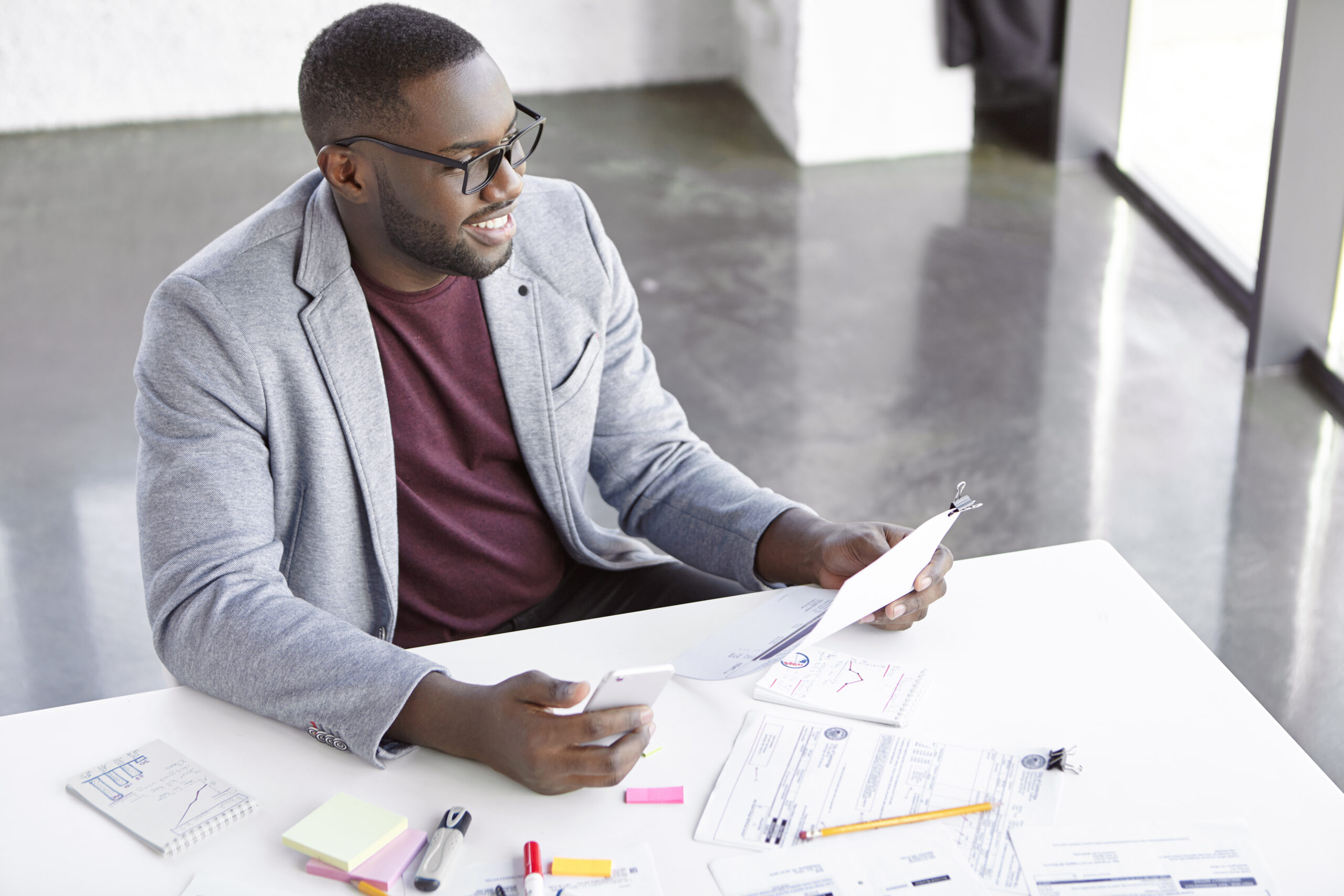 Indoor Shot Of Smiling Office Worker With Dark Skin Holds Document And Smart Phone, Downloads Online Popular Application Or Multimedia Program On Mobile Store, Happy To Have Successful Working Day