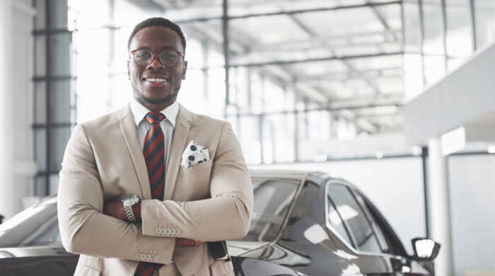 Young Black Businessman On Auto Salon Background. Car Sale And Rent Concept